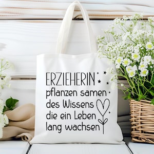 May include: White tote bag with black text that reads "ERZIEHERIN pflanzen samen des Wissens die ein Leben lang wachsen" with a heart and star design. The bag is on a white wooden surface, with white flowers in the background.