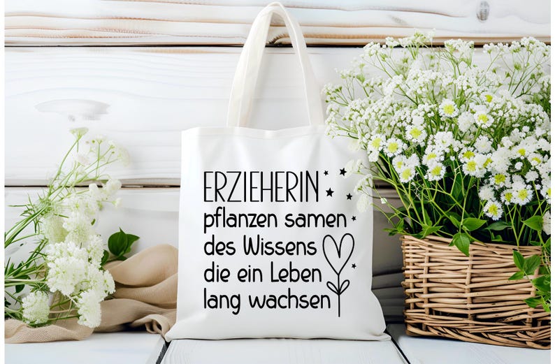 May include: White tote bag with black text that reads "ERZIEHERIN pflanzen samen des Wissens die ein Leben lang wachsen" with a heart and star design. The bag is on a white wooden surface, with white flowers in the background.