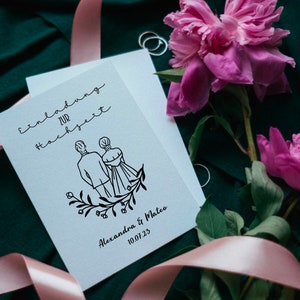 May include: A white wedding invitation with black calligraphy that reads "Einladung zur Hochzeit" and a simple line drawing of a couple holding hands. The text below the drawing reads "Alexandra & Mateo 10.07.23". The invitation is surrounded by pink peonies and a pink ribbon.