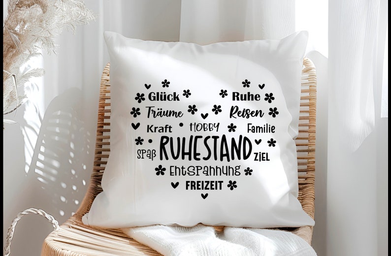 May include: White square pillow with a black heart-shaped design. The design includes the word "RUHESTAND" and other German words like "Gl&uuml;ck", "Tr&auml;ume", "Ruhe", "Reisen", "Familie", "Freizeit", and "Entspannung".