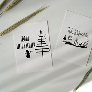 May include: Two white cards with black ink illustrations and the text "Frohe Weihnachten" on each. One card features a Christmas tree, snowman, and stars. The other card features a Christmas tree, houses, a deer, and hearts.