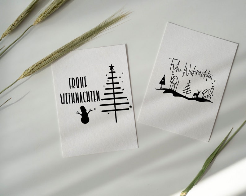 May include: Two white cards with black ink illustrations and the text "Frohe Weihnachten" on each. One card features a Christmas tree, snowman, and stars. The other card features a Christmas tree, houses, a deer, and hearts.