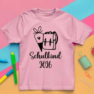 May include: A light pink t-shirt with a black graphic of a school backpack and a carrot. The text "Schulkind 2026" is printed below the graphic in a script font. The shirt is on a colorful background.