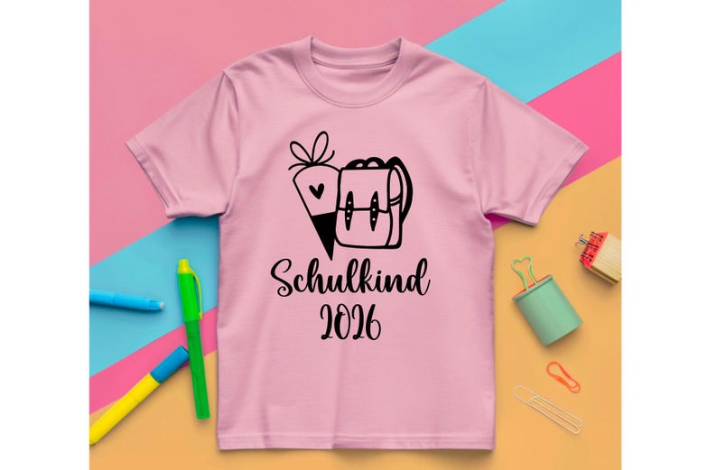 May include: A light pink t-shirt with a black graphic of a school backpack and a carrot. The text "Schulkind 2026" is printed below the graphic in a script font. The shirt is on a colorful background.