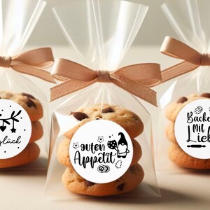 May include: Three clear cellophane bags tied with brown ribbon, each containing a stack of chocolate chip cookies with a round sticker on top. The stickers feature black and white illustrations and text in German. The stickers say "Keksögluck", "Guten Appetit", and "Backen mit Liebe".