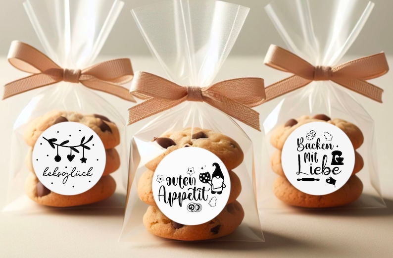 May include: Three clear cellophane bags tied with brown ribbon, each containing a stack of chocolate chip cookies with a round sticker on top. The stickers feature black and white illustrations and text in German. The stickers say "Keksögluck", "Guten Appetit", and "Backen mit Liebe".