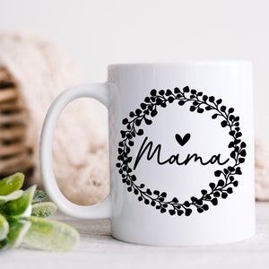 May include: White ceramic mug with a black floral wreath design and the word "Mama" inside the wreath. A small black heart is above the word "Mama".