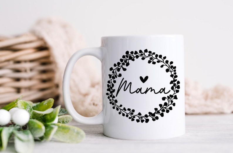 May include: White ceramic mug with a black floral wreath design and the word "Mama" inside the wreath. A small black heart is above the word "Mama".