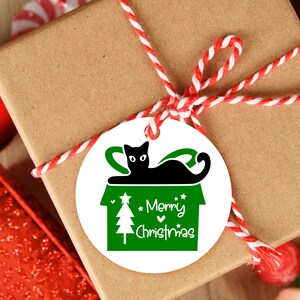 May include: A white round gift tag with a green and black illustration of a cat sitting in a house with a Christmas tree. The text "Merry Christmas" is written in green on the tag.