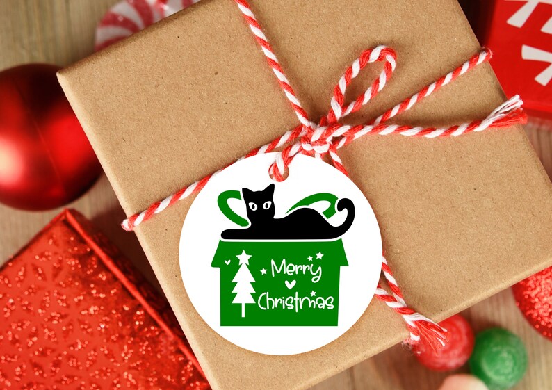 May include: A white round gift tag with a green and black illustration of a cat sitting in a house with a Christmas tree. The text "Merry Christmas" is written in green on the tag.