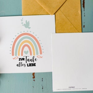 May include: A white card with a rainbow, a dove, and the text "ZUR Taufe alles LIEBE" in black. The card is on a blue wooden surface with a brown envelope and purple flowers.
