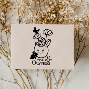 May include: A white card with a black and white illustration of a bunny in an egg-shaped pot with flowers and a butterfly. The text "Gruss vom Osterhasi" is below the illustration.