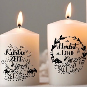 May include: Two white pillar candles with black text and illustrations. The candle on the left says "K&uuml;rbis Zeit" with a pumpkin and mushrooms. The candle on the right says "Herbst Liebe" with a wreath of leaves and pumpkins.