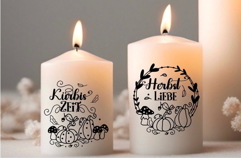 May include: Two white pillar candles with black text and illustrations. The candle on the left says "K&uuml;rbis Zeit" with a pumpkin and mushrooms. The candle on the right says "Herbst Liebe" with a wreath of leaves and pumpkins.