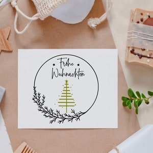 May include: A white card with black text that reads "Frohe Weihnachten" and a stylised green Christmas tree with gold stars inside a black circle with a black branch design.