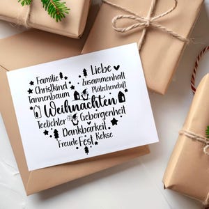 May include: A white card with black text and illustrations of houses and stars. The text reads "Familie, Christkind, Tannenbaum, Teelichter, Geborgenheit, Dankbarkeit, Freude Fest Kekse, Liebe, Zusammenhall, Pl&auml;tzchenduf", and "Weihnachten".