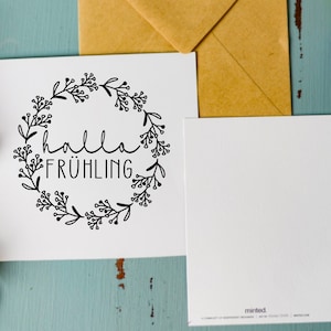 May include: A white card with a black floral wreath and the text "hallo fr&uuml;hling" inside the wreath. The card is on a blue wooden surface with a brown envelope and purple flowers.