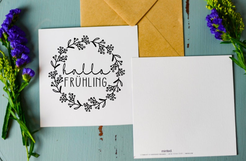 May include: A white card with a black floral wreath and the text "hallo fr&uuml;hling" inside the wreath. The card is on a blue wooden surface with a brown envelope and purple flowers.