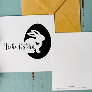 May include: A white card with a black circle containing a white silhouette of a rabbit with a heart. The text "Frohe Ostern" is written in black cursive script below the circle.