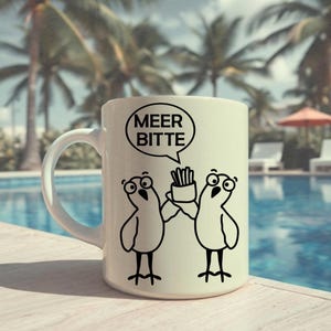 May include: White ceramic mug with a cartoon illustration of two birds and the text "MEER BITTE" in a speech bubble. One bird is holding fries. The mug is placed near a swimming pool with palm trees in the background.