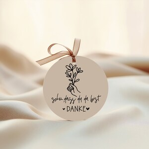 Peut inclure: Une &eacute;tiquette ronde de couleur cr&egrave;me avec une illustration en noir et blanc d'une main tenant un bouquet de fleurs. L'&eacute;tiquette porte le texte "sch&ouml;n dass du da bist &hearts; DANKE &hearts;" imprim&eacute; dessus.