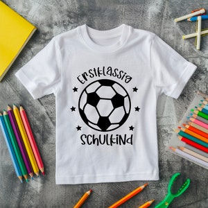May include: White t-shirt with a black and white soccer ball graphic and the text "Erstklassig SCHULKIND". The shirt is surrounded by colored pencils and a yellow notebook.