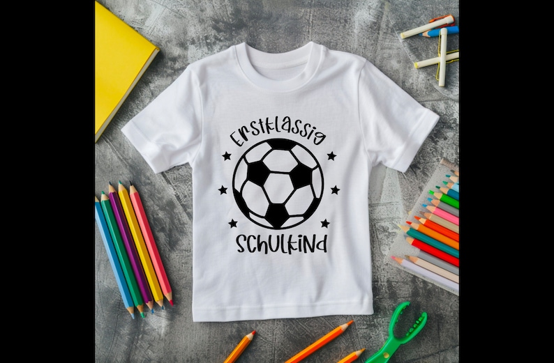 May include: White t-shirt with a black and white soccer ball graphic and the text "Erstklassig SCHULKIND". The shirt is surrounded by colored pencils and a yellow notebook.