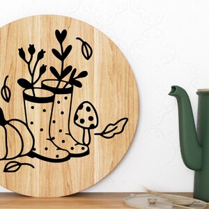 Op de afbeelding: Een rond houten bord met een zwart-witte illustratie van een pompoen, twee regenlaarzen met stippen, een paddenstoel en bladeren. De laarzen zijn gevuld met bloemen.