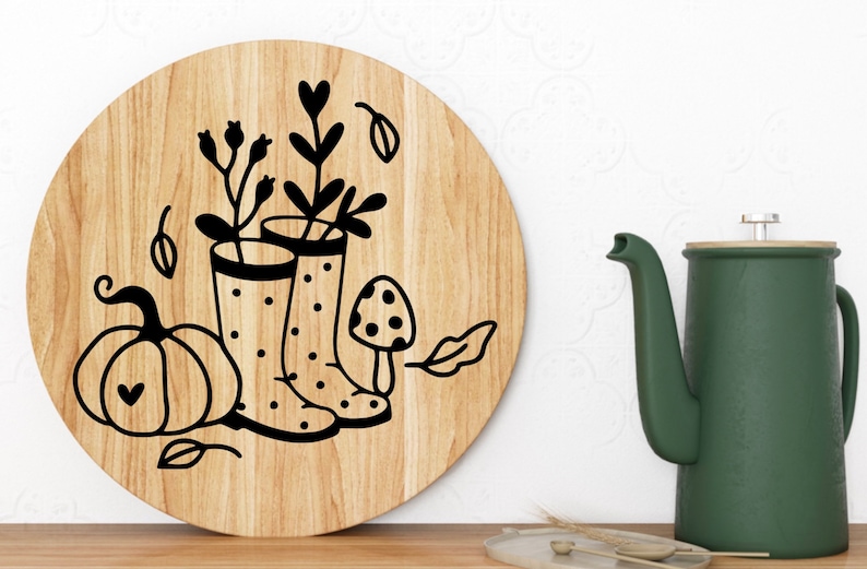 Op de afbeelding: Een rond houten bord met een zwart-witte illustratie van een pompoen, twee regenlaarzen met stippen, een paddenstoel en bladeren. De laarzen zijn gevuld met bloemen.