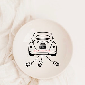 Op de afbeelding: Een witte keramische ringenschotel met een zwarte lijntekening van een auto met de woorden "LOVE" en "just married" op de achterkant. De auto heeft aan elke kant een hart en is vastgebonden met een lint.