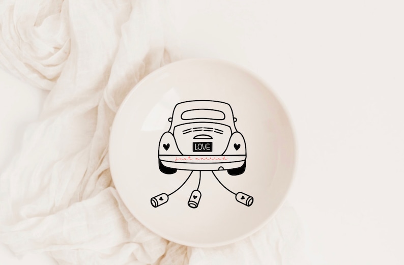 Op de afbeelding: Een witte keramische ringenschotel met een zwarte lijntekening van een auto met de woorden "LOVE" en "just married" op de achterkant. De auto heeft aan elke kant een hart en is vastgebonden met een lint.
