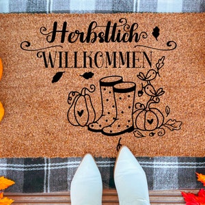 May include: A brown coir doormat with the text "Herbstlich Willkommen" in black lettering. The doormat features a pair of black rain boots with white polka dots and a fall-themed design with pumpkins and leaves.
