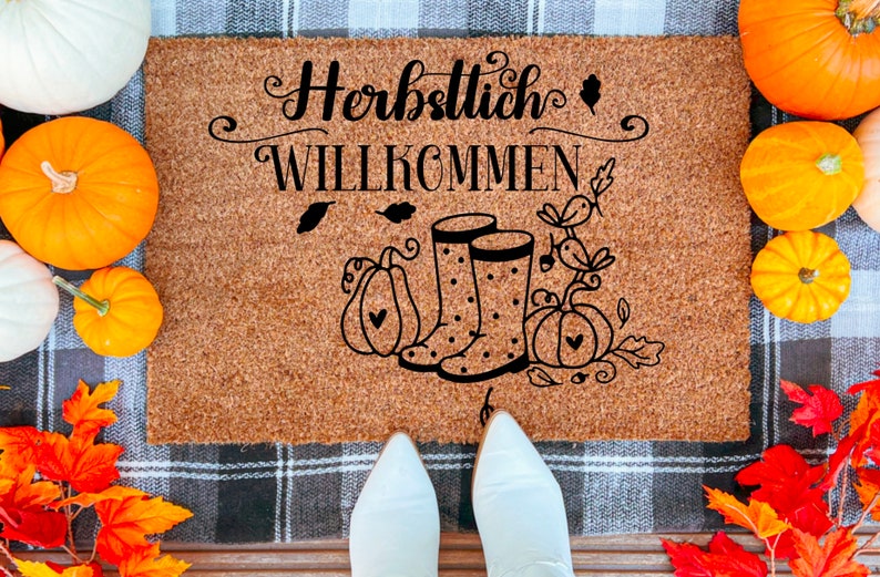 May include: A brown coir doormat with the text "Herbstlich Willkommen" in black lettering. The doormat features a pair of black rain boots with white polka dots and a fall-themed design with pumpkins and leaves.
