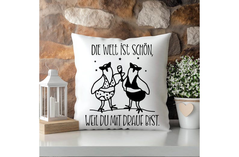 May include: White square cushion featuring a black illustration of two birds, one holding a glass. The cushion has the German text "DIE WELT IST SCH&Ouml;N, WEIL DU MIT DRAUF BIST." Decorative cushion for home decor.