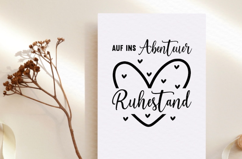 May include: A white card with black text that reads "AUF INS Abenteuer Ruhestand" in a decorative font, with a heart-shaped design. A dried plant branch is in the background.