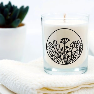May include: A white candle in a clear glass jar with a black floral design. The candle is sitting on a white towel.