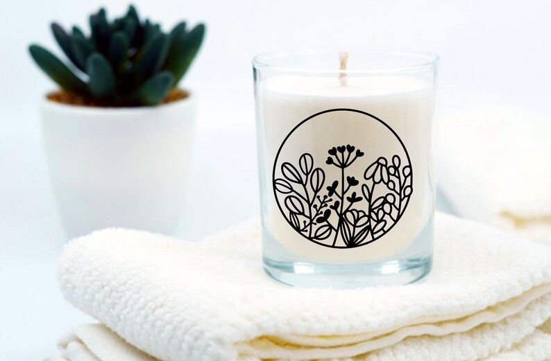 May include: A white candle in a clear glass jar with a black floral design. The candle is sitting on a white towel.