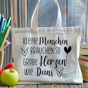 May include: A white canvas tote bag with black lettering that reads "KLEINE Menschen BRAUCHEN SO GROBE Herzen Wie Deins" and heart illustrations. The bag is on a wooden surface with pencils, apples, and books.