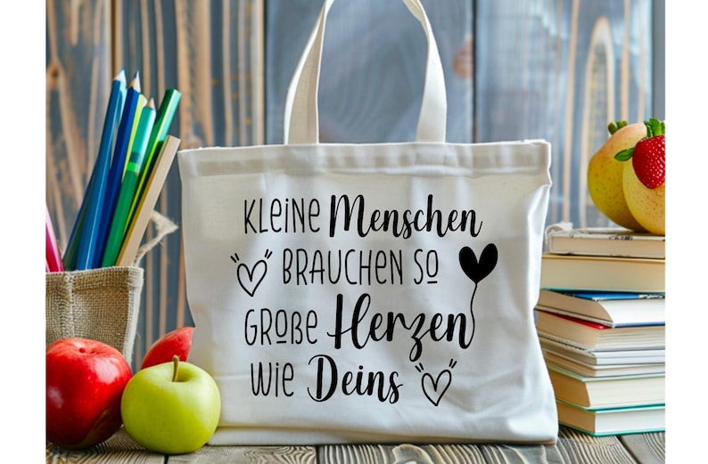 May include: A white canvas tote bag with black lettering that reads "KLEINE Menschen BRAUCHEN SO GROBE Herzen Wie Deins" and heart illustrations. The bag is on a wooden surface with pencils, apples, and books.