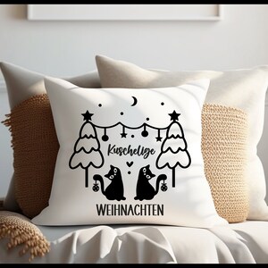 May include: White throw pillow with black text and illustrations. The text reads "Kuschelige Weihnachten" which translates to "Cozy Christmas" in English. The illustration features two black cats sitting under a string of lights between two Christmas trees.