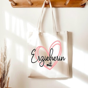 May include: A cream-colored tote bag with the German text "Erzieherin mit" inside a pink heart design. The bag hangs from a wooden rack against a white wall, with dried plants in the foreground.