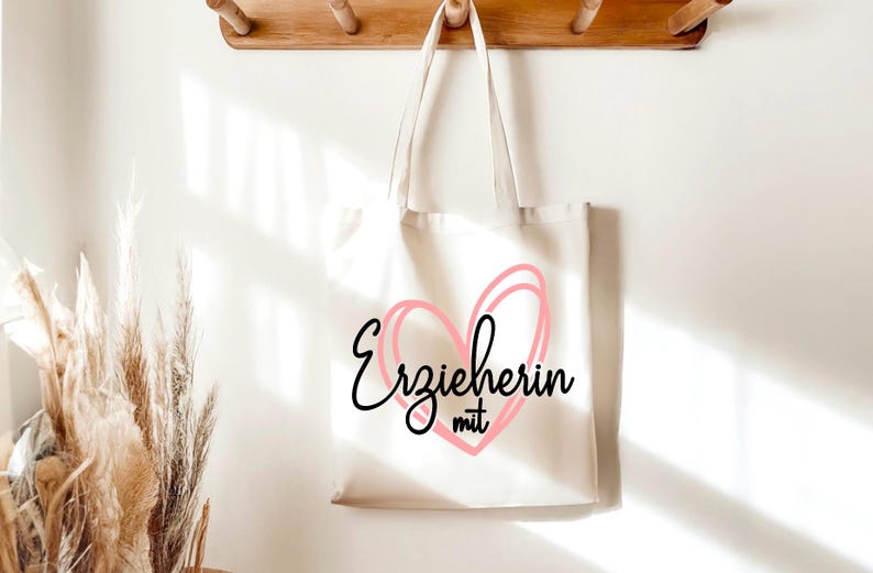 May include: A cream-colored tote bag with the German text "Erzieherin mit" inside a pink heart design. The bag hangs from a wooden rack against a white wall, with dried plants in the foreground.