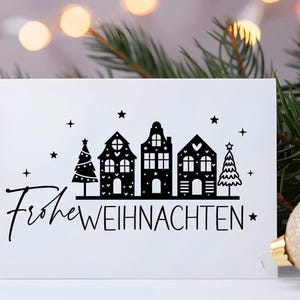 Puede incluir: Una tarjeta blanca con texto negro e ilustraciones. El texto dice "Frohe Weihnachten" y las ilustraciones son de una fila de casas con corazones y estrellas, y dos &aacute;rboles de Navidad.