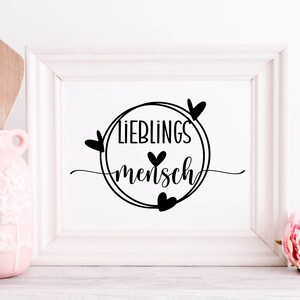 May include: A white framed print with black text that reads "Lieblingsmensch" in a circle with hearts around it.