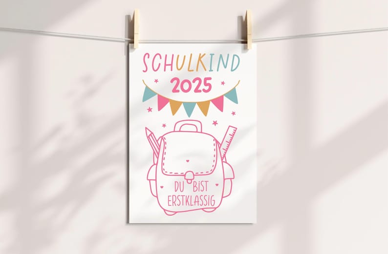 May include: A white poster with a pink outline illustration of a backpack, pencil, and ruler. The text "SCHULKIND 2025" is at the top, with the words "DU BIST ERSTKLASSIG" at the bottom. The poster is held by wooden clothespins.