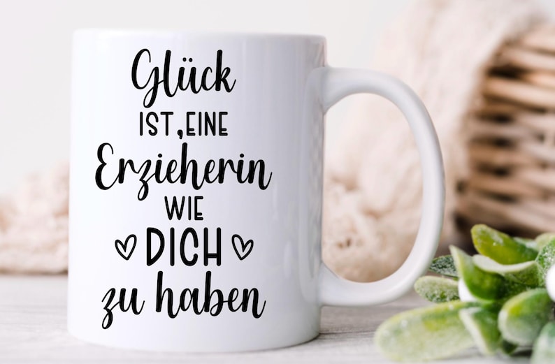 May include: White ceramic mug with a black inscription in German: "Gl&uuml;ck ist, eine Erzieherin wie DICH zu haben" (Happiness is having a teacher like YOU). The mug has a curved handle and is set against a blurred background.