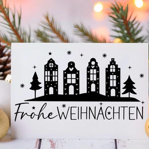 Puede incluir: Una tarjeta blanca con una silueta negra de una fila de casas con un &aacute;rbol de Navidad a cada lado. El texto "Frohe Weihnachten" est&aacute; escrito debajo de las casas.