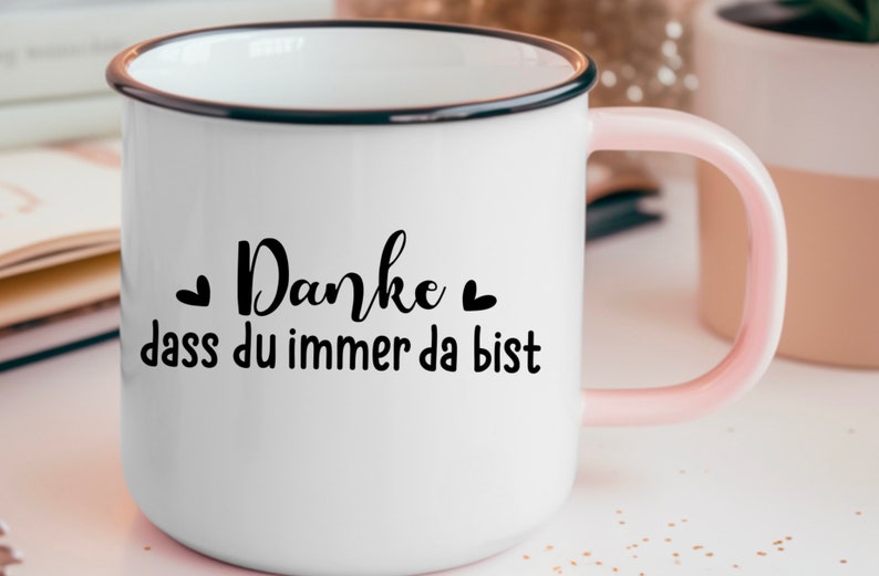 Puede incluir: Taza de esmalte blanco con borde negro y asa rosa. La taza tiene texto negro que dice "Danke dass du immer da bist" con un peque&ntilde;o coraz&oacute;n a cada lado de la palabra "Danke".
