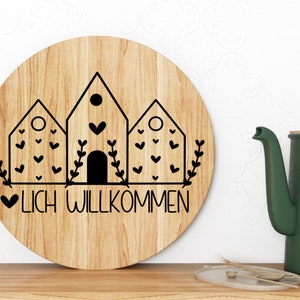 May include: A round wooden sign with a black and white design of three houses with hearts and greenery. The sign says "LICH WILLKOMMEN" in black lettering.