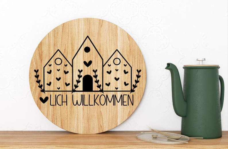 May include: A round wooden sign with a black and white design of three houses with hearts and greenery. The sign says "LICH WILLKOMMEN" in black lettering.
